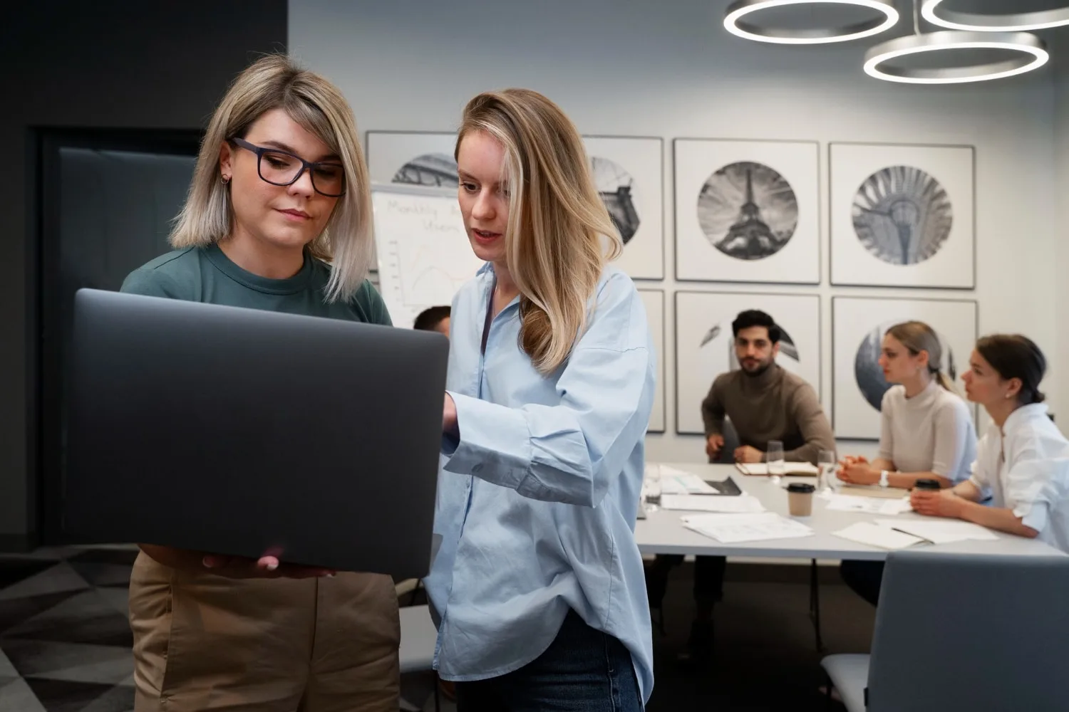 Two professionals collaborating on a laptop in a modern conference room, while a team discusses data in the background, representing the effectiveness of scalable IT teams in driving business success and innovation.