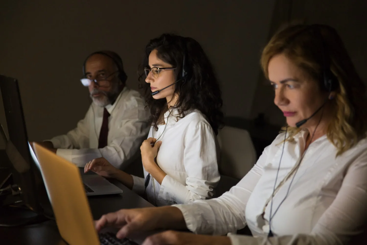 A team of customer support professionals wearing headsets and working on laptops in a dimly lit office, illustrating the efficiency of business IT support through skilled remote staffing solutions.