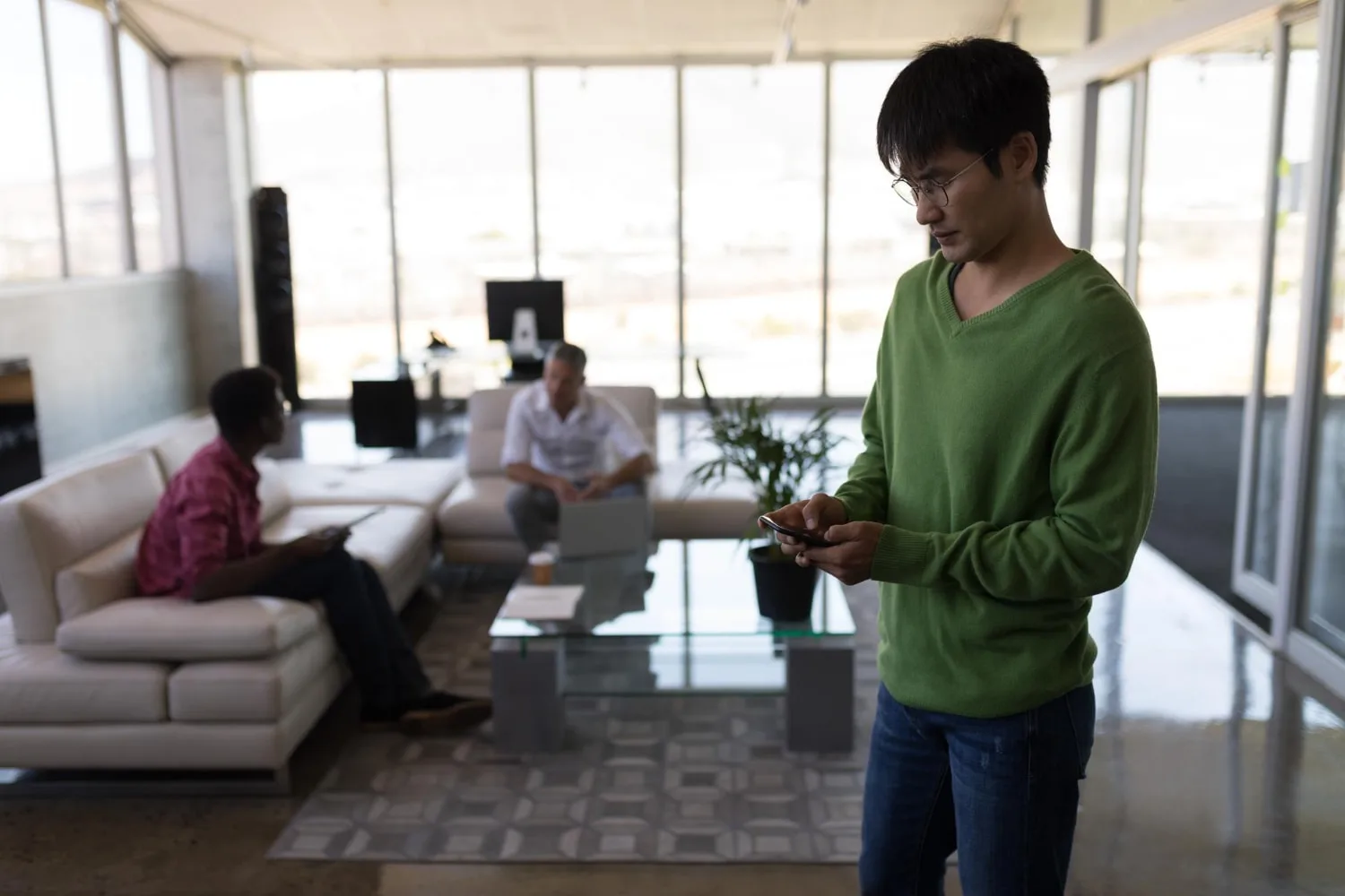 A professional in a green sweater focuses on his phone while two colleagues have a discussion in a modern, well-lit office lounge, representing the flexibility and efficiency of tech talent outsourcing.